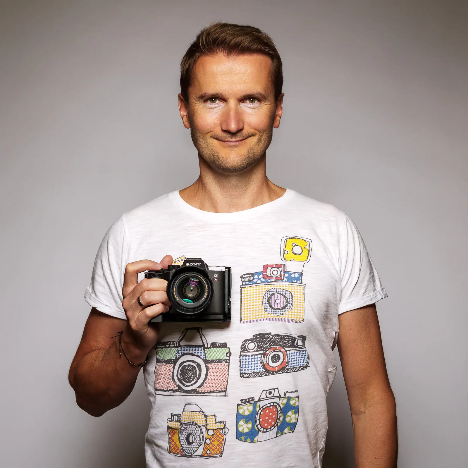 Smiling man with light brown hair holding a Sony camera in front of his chest, wearing a white t-shirt printed with colorful retro camera illustrations on a plain light background.